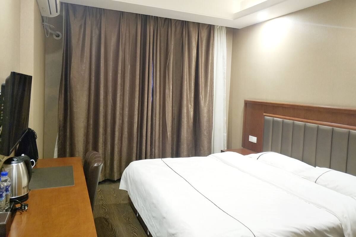 A hotel room with a bed, a desk with a kettle, a chair, and a curtain.