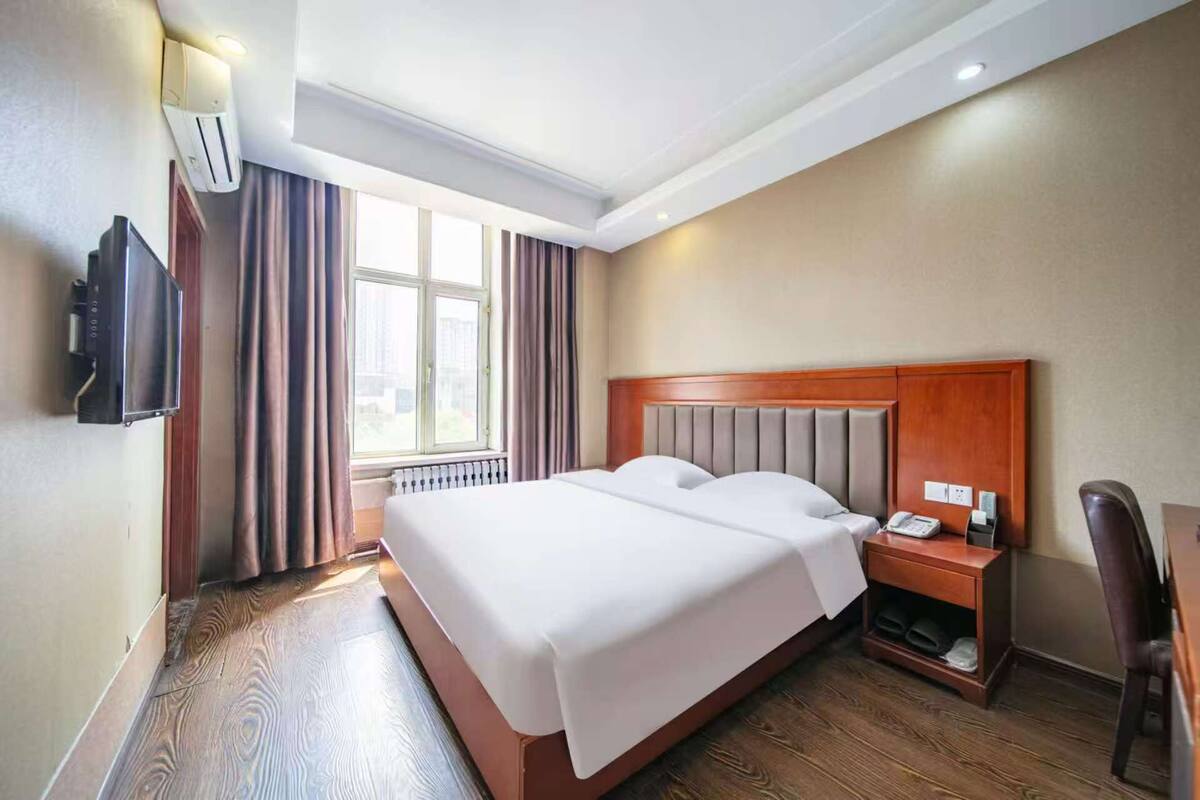 A hotel room with a large bed, a bedside table with a phone, a TV mounted on the wall, and a window with curtains.