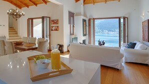 Een ruime woonkamer met een hoge plafond en houten balken, een eettafel met een schaal fruit, en een groot raam met uitzicht op de zee.