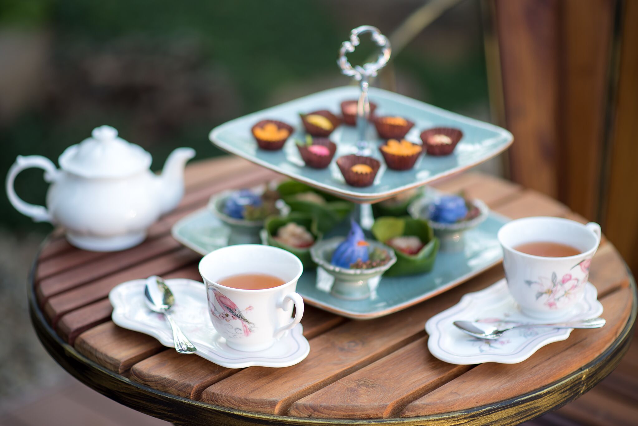A tea set on a round wooden table with a teapot, two cups, and a tiered tray with cupcakes.