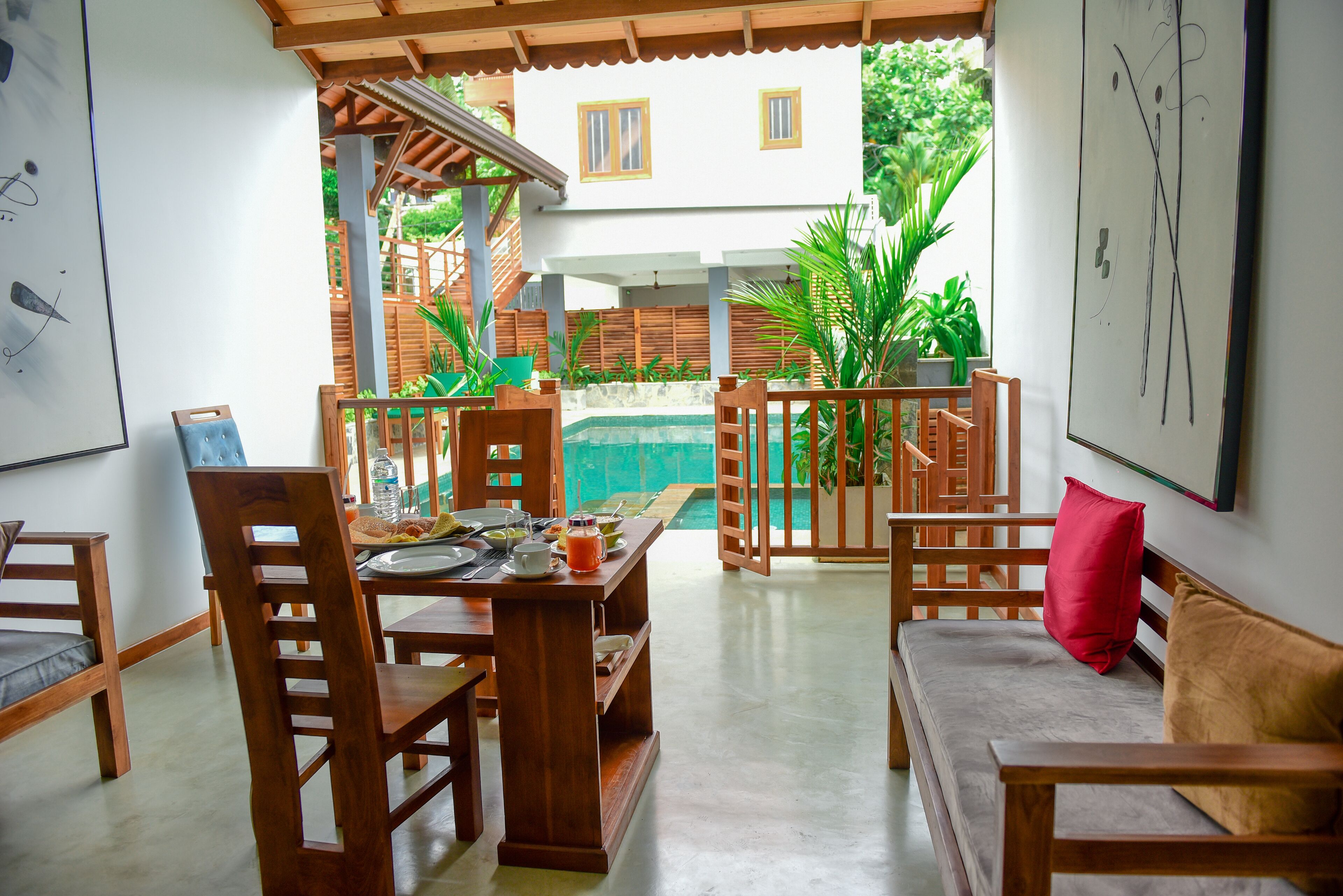 A dining area with wooden furniture, a table set with plates, glasses, and a fruit bowl. There is a pool visible through the glass doors in the background.