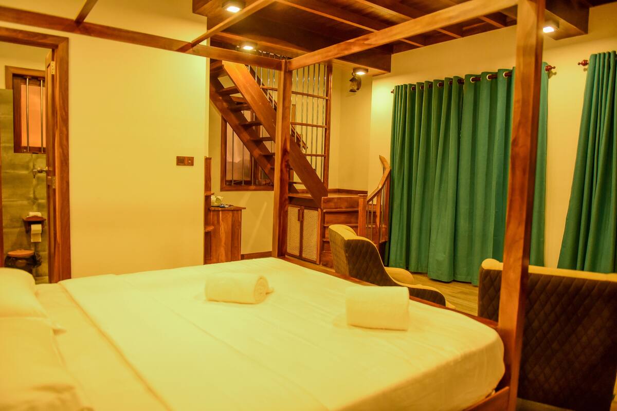 A hotel room with a wooden staircase, a bed with two pillows, a chair, and a green curtain.