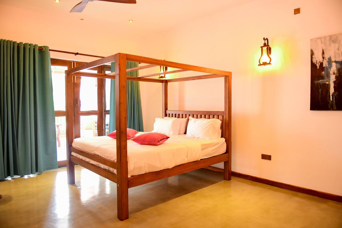 A four-poster bed with white bedding and red pillows, a ceiling fan, and a wall-mounted light fixture.