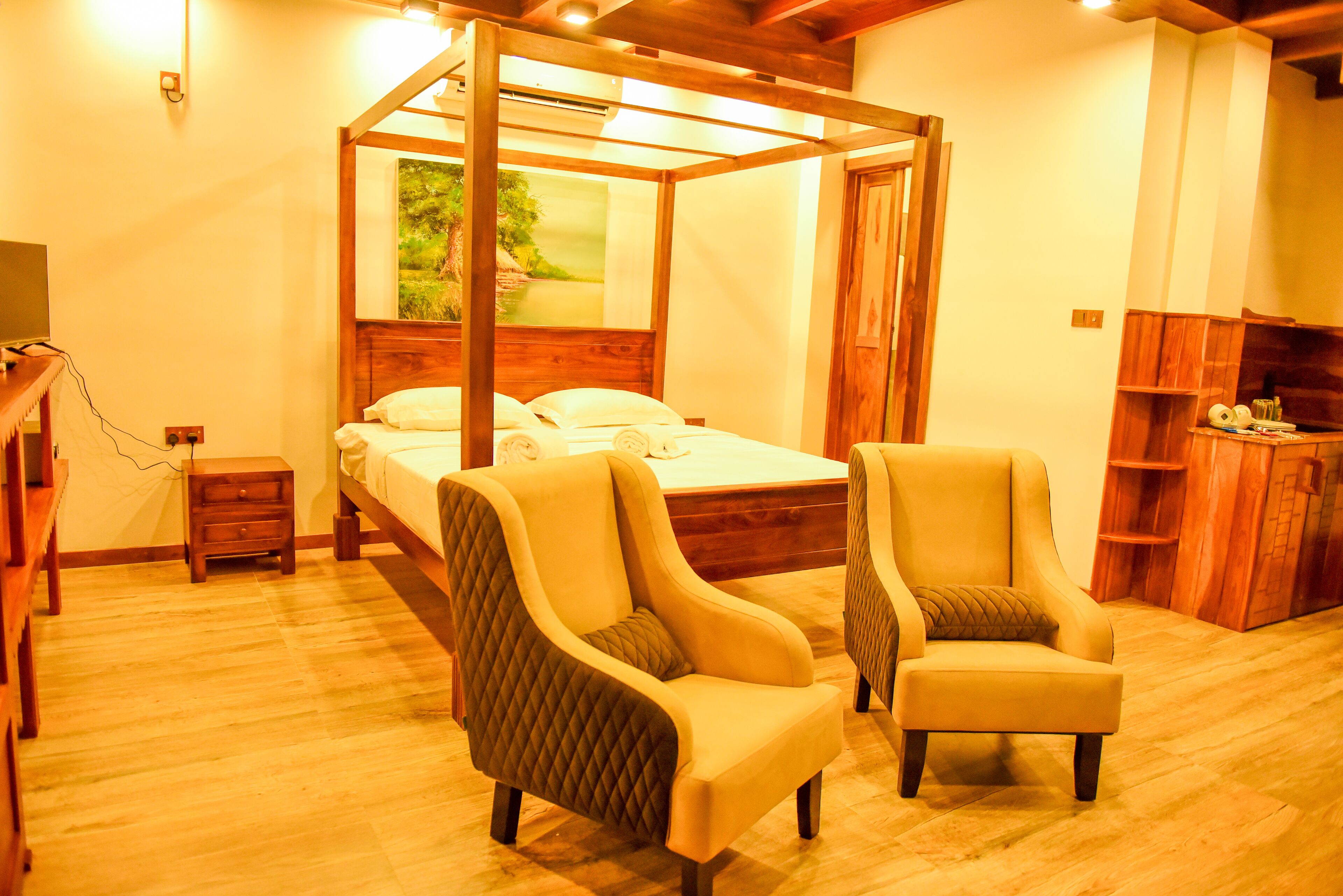 A four-poster bed with a wooden headboard, two armchairs with cushions, a small bedside table, and a wooden shelf unit.