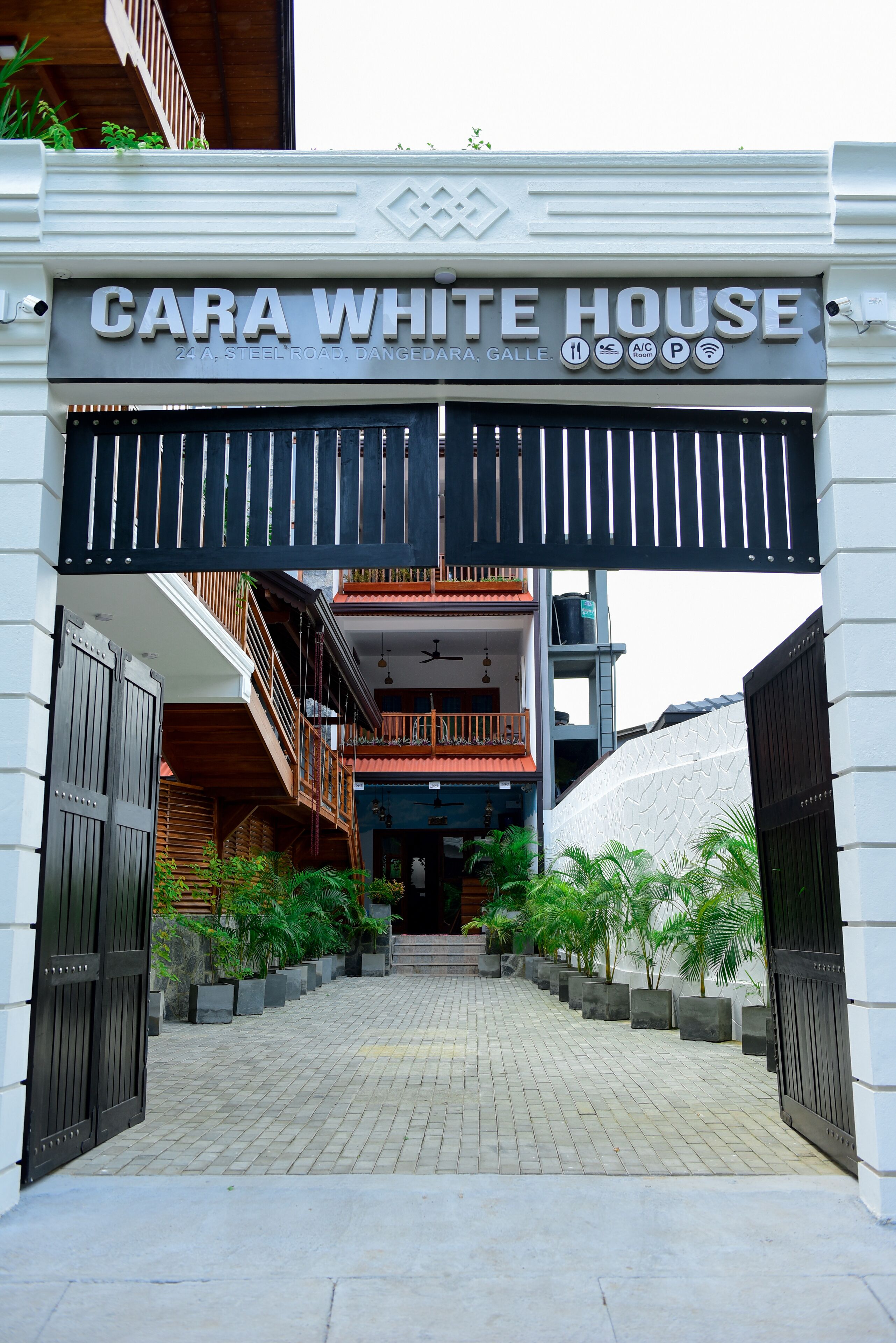 The entrance to Cara White House, featuring a gated driveway, potted plants, and a signboard with amenities.