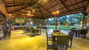 Un área de patio techada al aire libre con mesas y sillas, un césped y árboles al fondo.