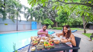 Un área de comedor junto a la piscina con una mesa para dos, rodeada de abundante vegetación y una piscina azul cristalina.