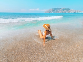 A woman sitting on a pebble beach with clear turquoise water and a distant hill.
