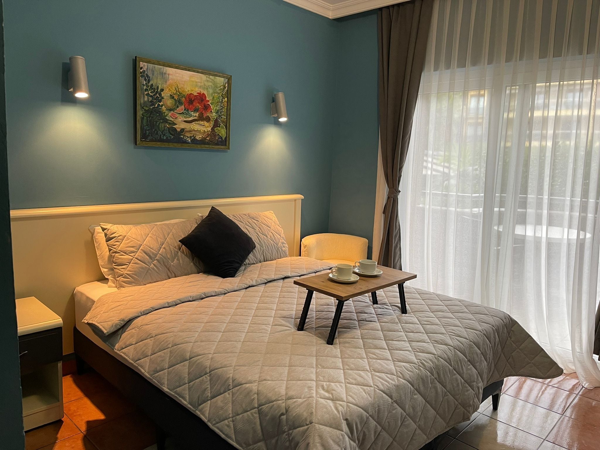 Un dormitorio con cama, mesitas de noche, un cuadro y una ventana con cortinas.