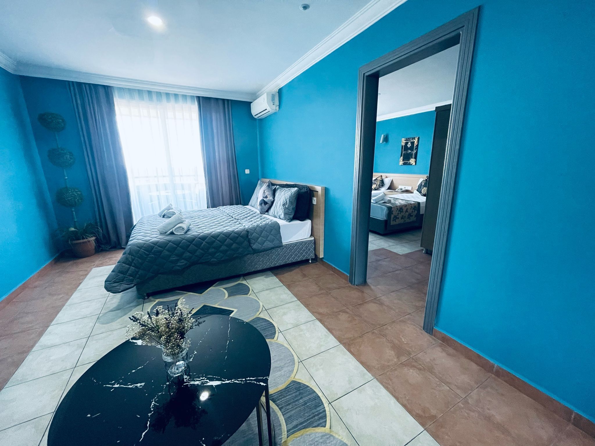 A hotel room with a bed, a small table with a vase, and a view of another room through an open door.