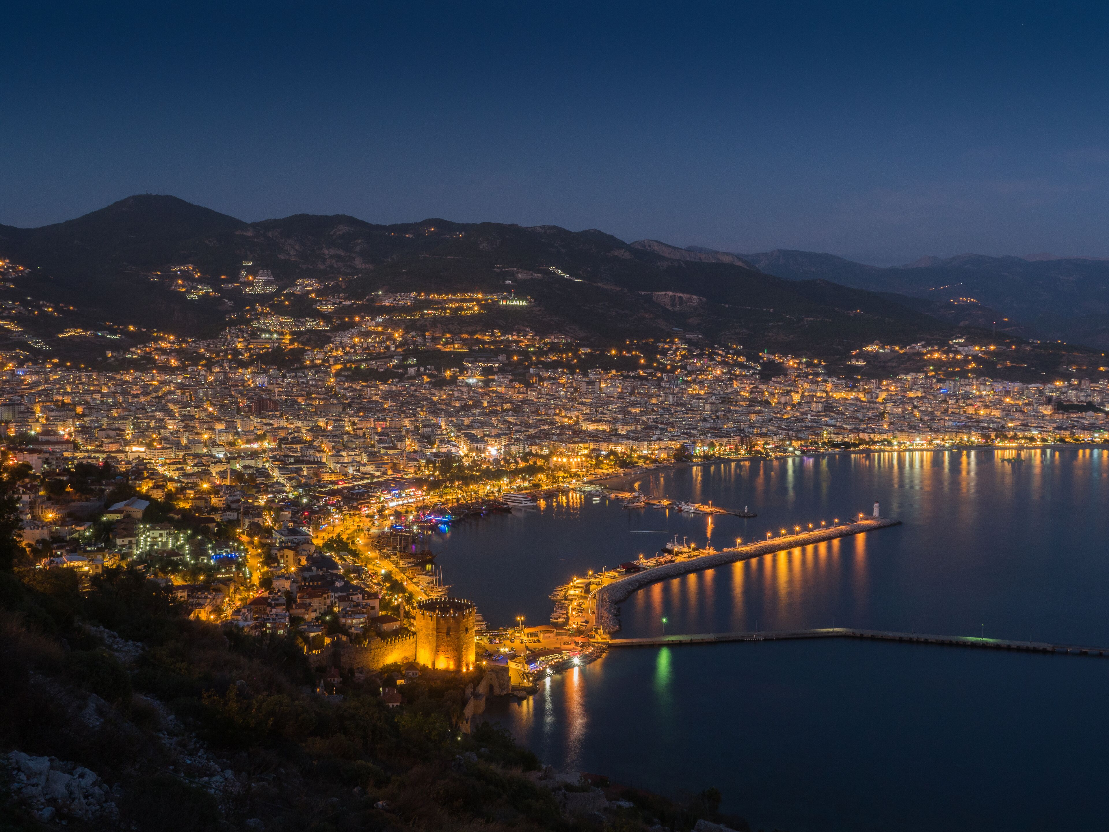 A coastal cityscape at night with a prominent castle on a hill, a marina, and a calm sea reflecting city lights.