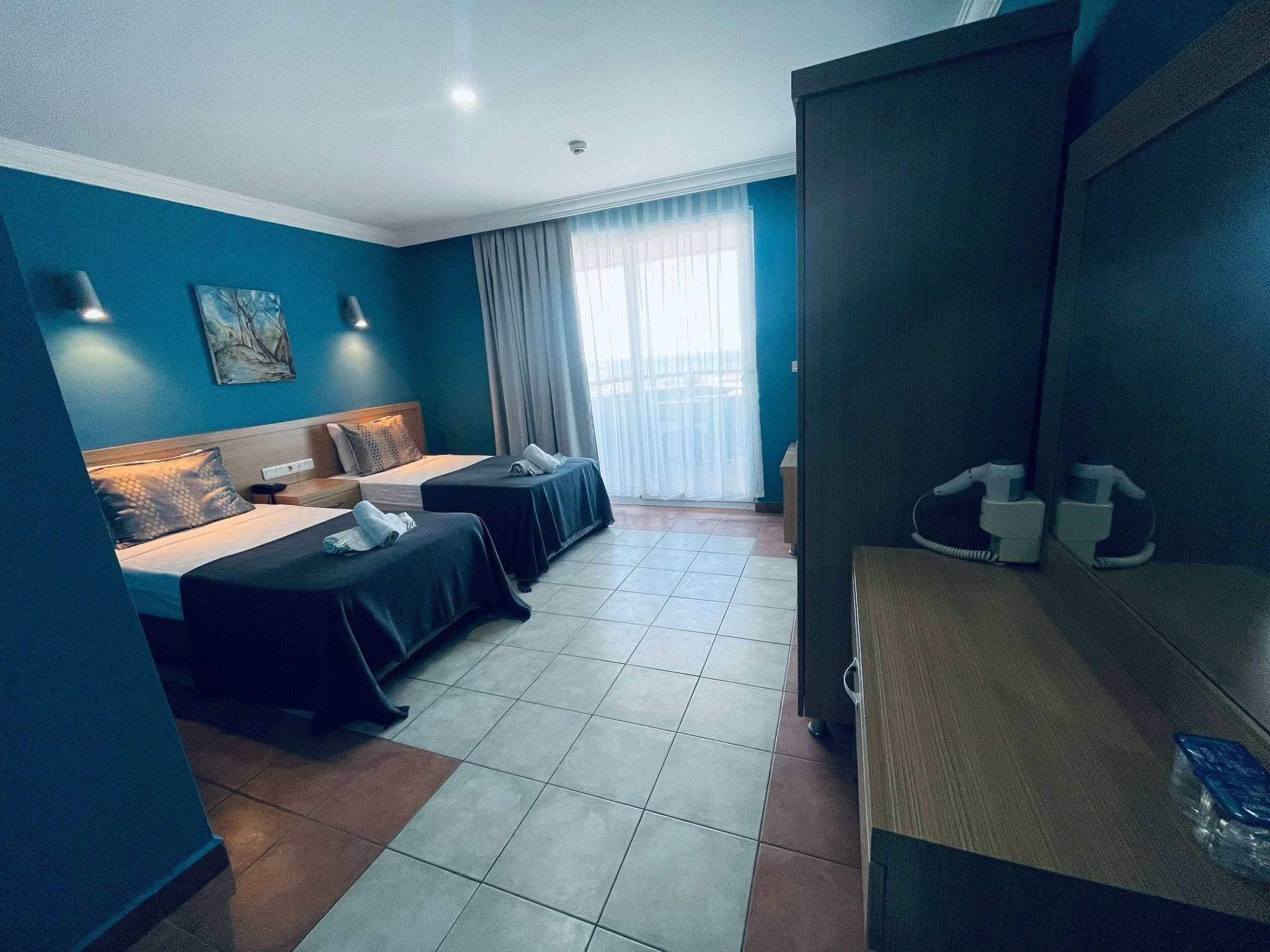 Habitación de hotel con dos camas, un sofá, un ventanal grande con cortinas y una mesita con una botella de agua.