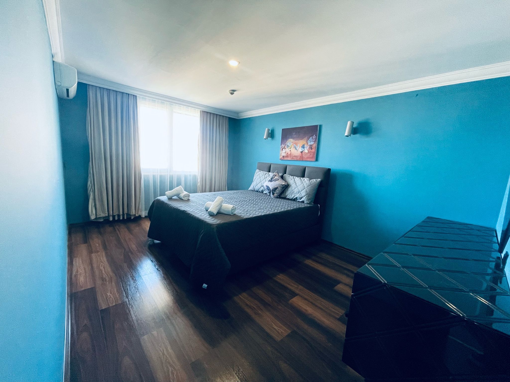 Quarto com cama, janela com cortinas, pintura na parede e piso de madeira.