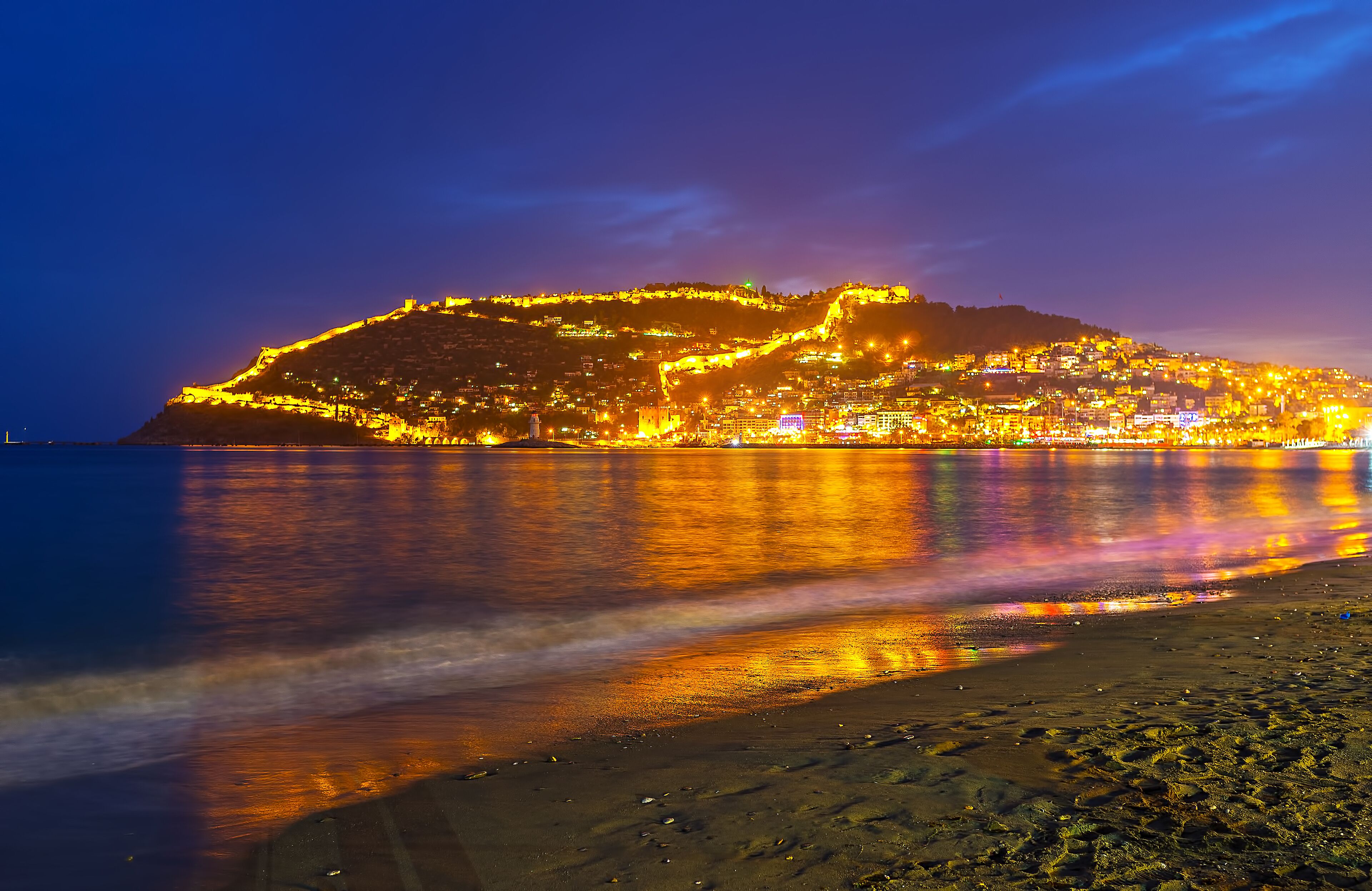 A coastal cityscape at night with illuminated buildings and a hilltop fortress.