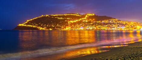 A coastal cityscape at night with illuminated buildings and a hilltop fortress.