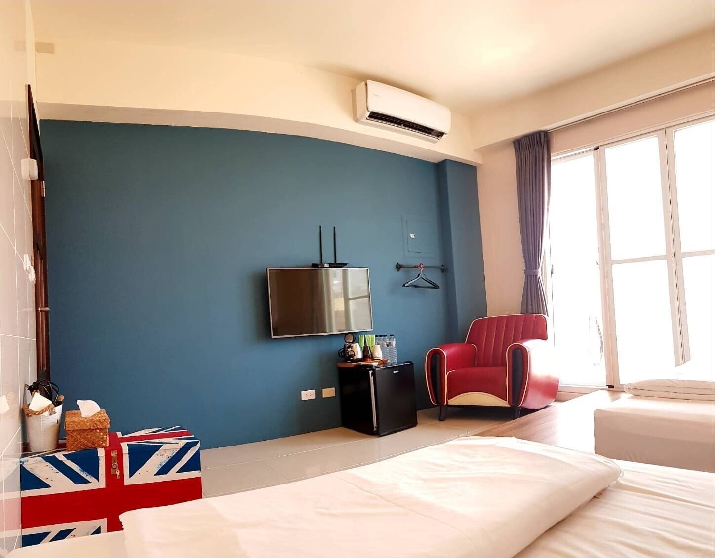 A hotel room with a bed, a TV mounted on the wall, a red chair, a small table with a lamp, and a window with curtains.