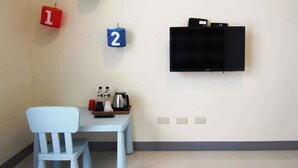 Una habitación con una silla blanca, una mesita pequeña con una tetera y tazas, y un televisor de pantalla plana montado en la pared. Hay paneles numerados en la pared.
