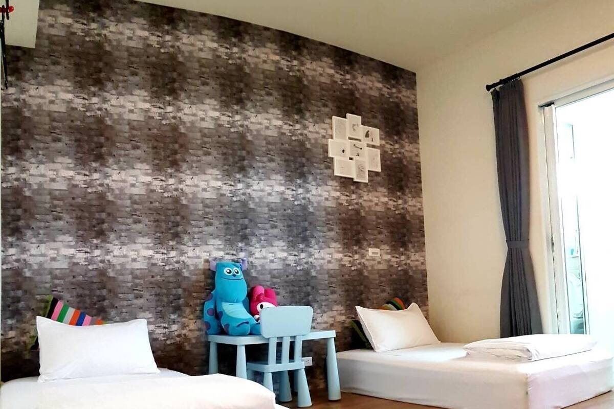 Una habitación de hotel con dos camas blancas, suelo de madera, pared con diseño de ladrillo, una mesa pequeña con una silla azul y una ventana con cortinas.