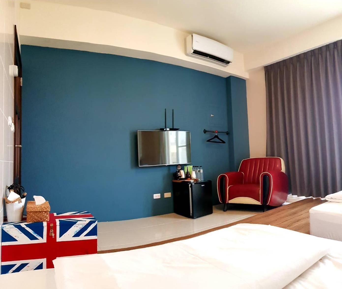 A hotel room with a bed, a red armchair, a flat-screen TV mounted on the wall, a small black side table, and a window with curtains.