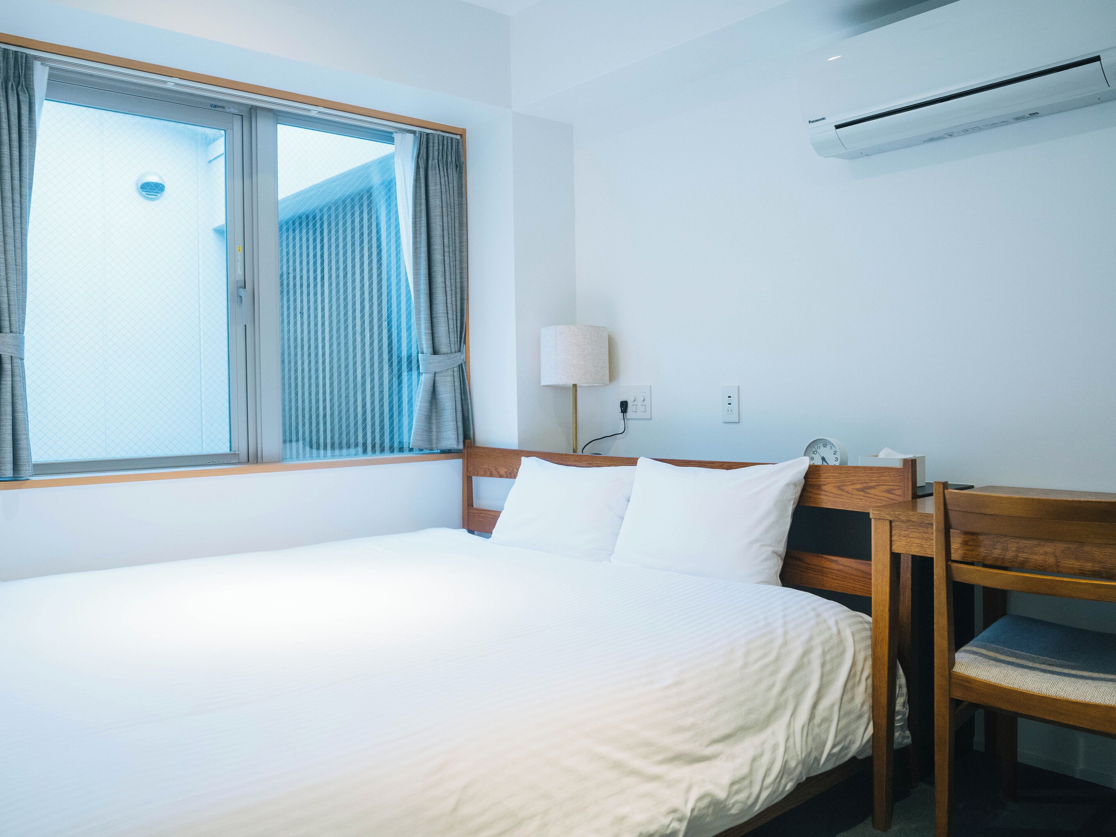 Habitación de hotel con una cama, un escritorio, una silla, una lámpara y un equipo de aire acondicionado.