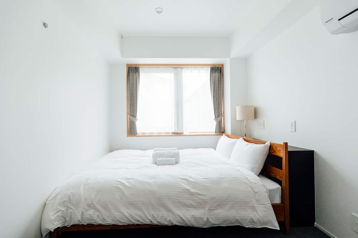 A neatly made bed with white linens and a wooden headboard in a bright room with a large window and a wall-mounted air conditioner.