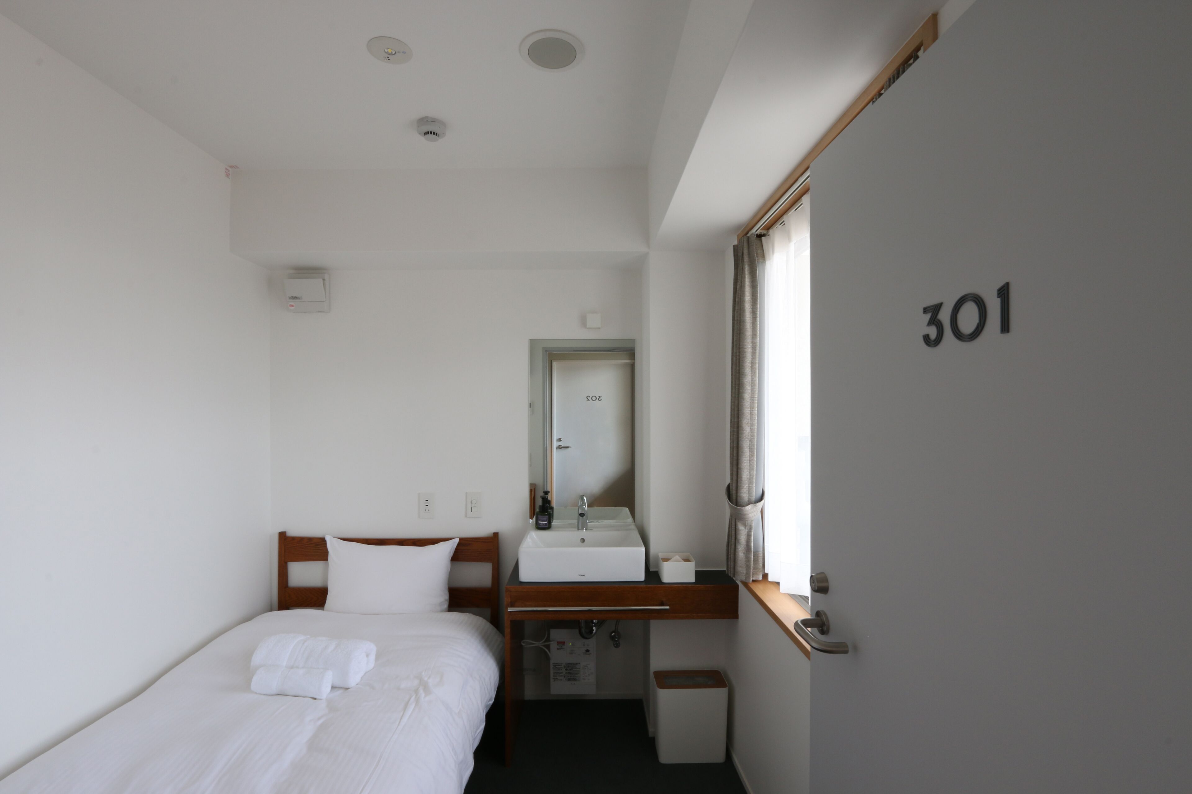 A small, clean room with a bed, a bedside table, a mirror, and a window. The room number is 301.