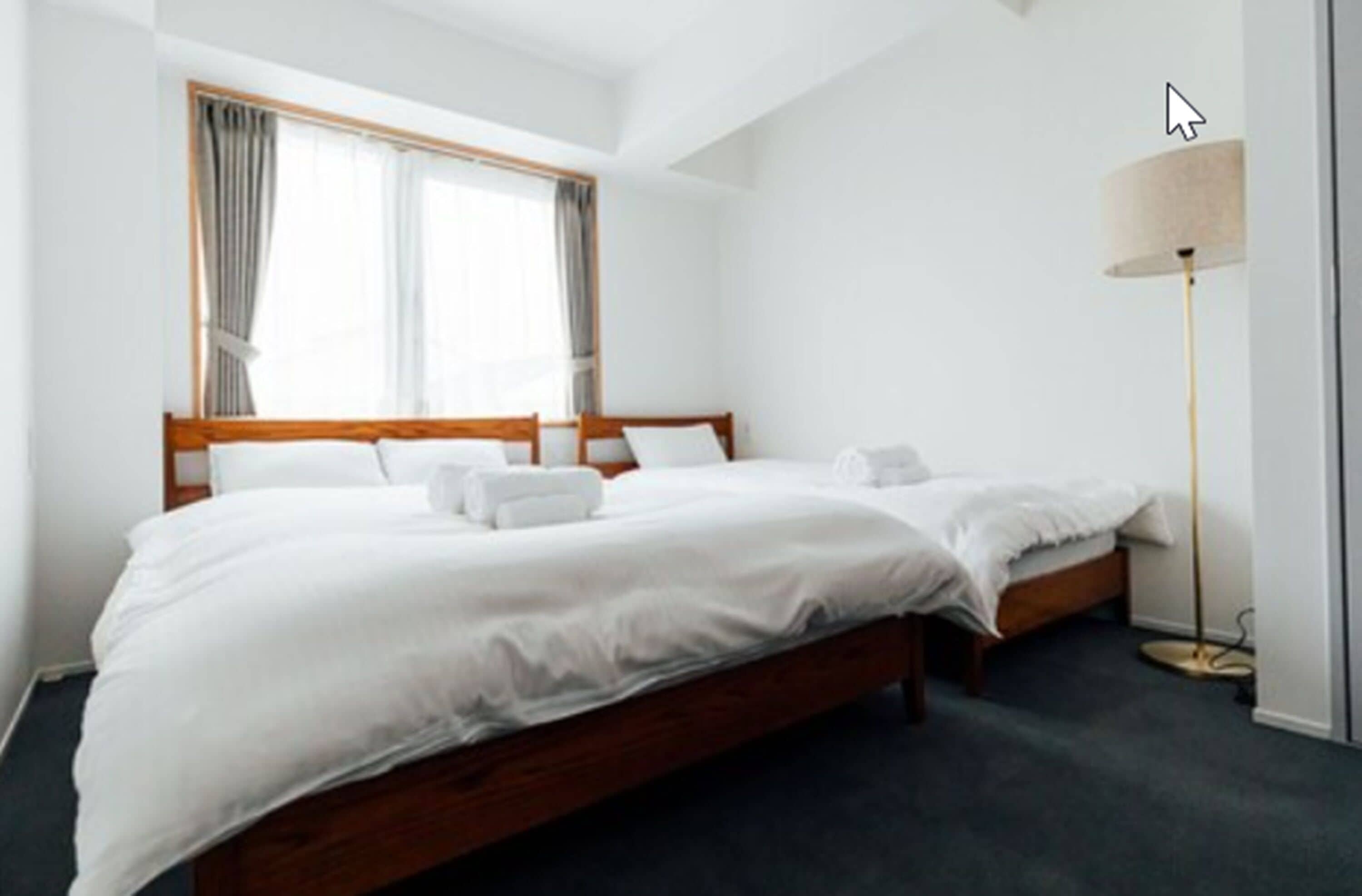A hotel room with two single beds, a large window with curtains, a standing floor lamp, and a dark carpeted floor.