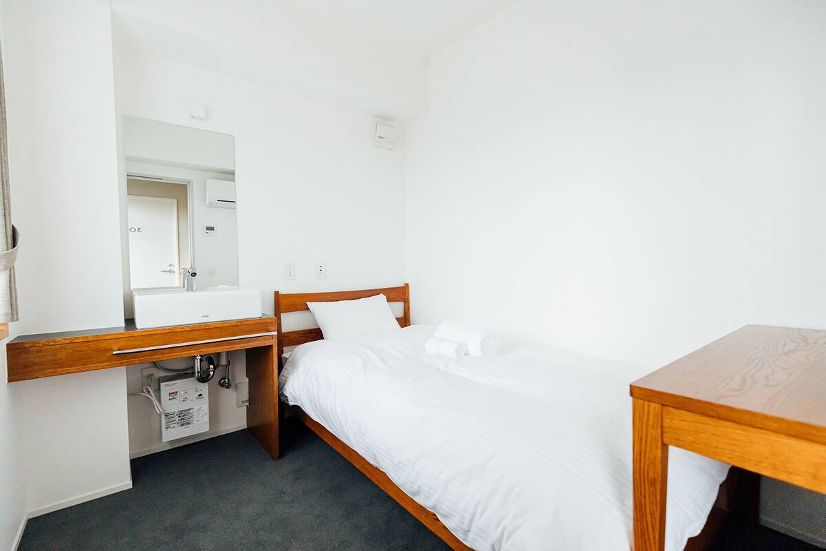 A small, clean room with a bed, a wooden nightstand, a mirror, and a white sink.