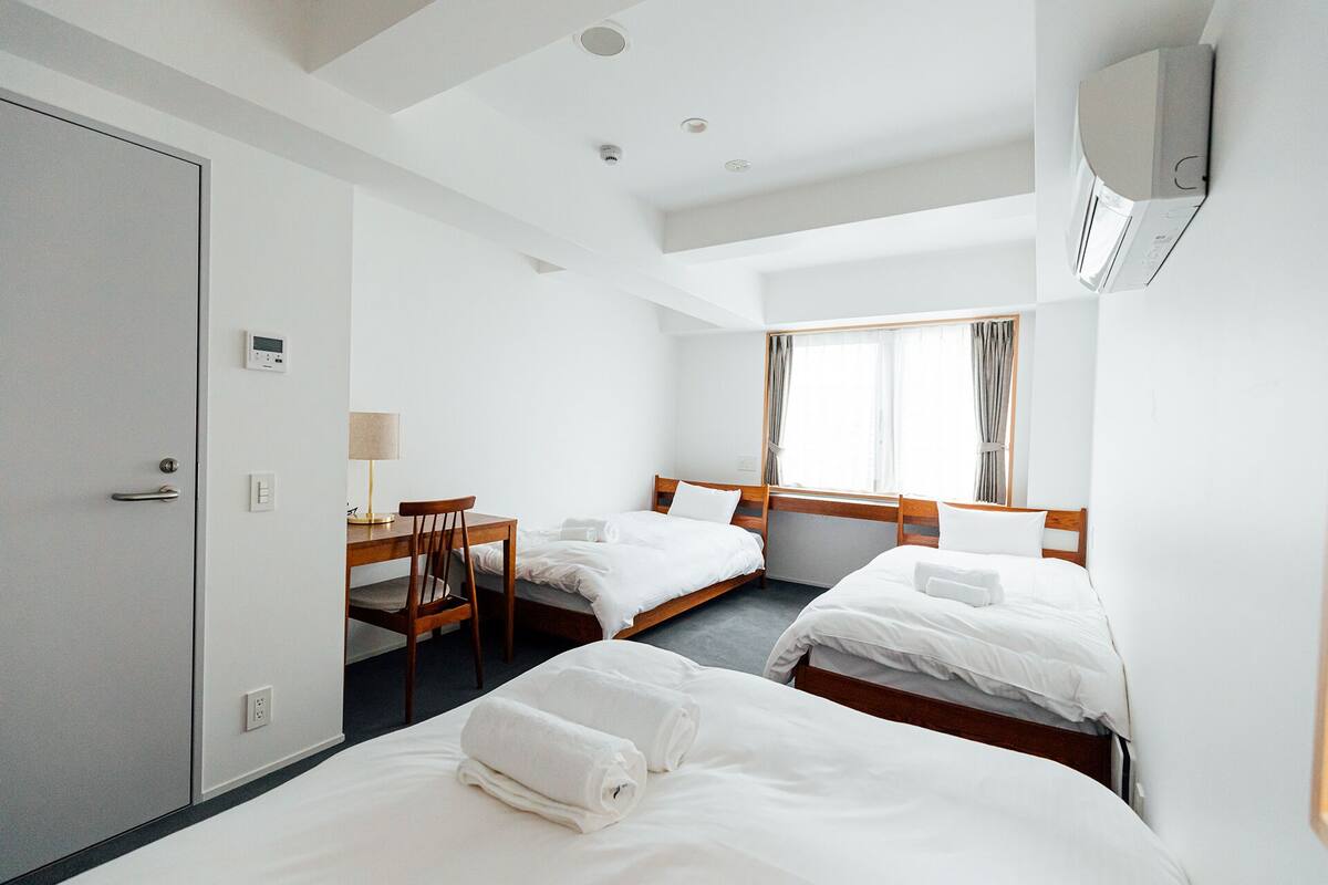 A hotel room with two beds, a desk, and a chair. There is a window with curtains, a door, and an air conditioning unit on the wall.