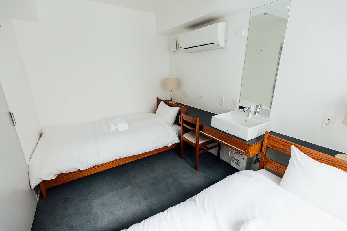 A small, clean room with two single beds, a wooden chair, a desk, a white sink, and a mirror.