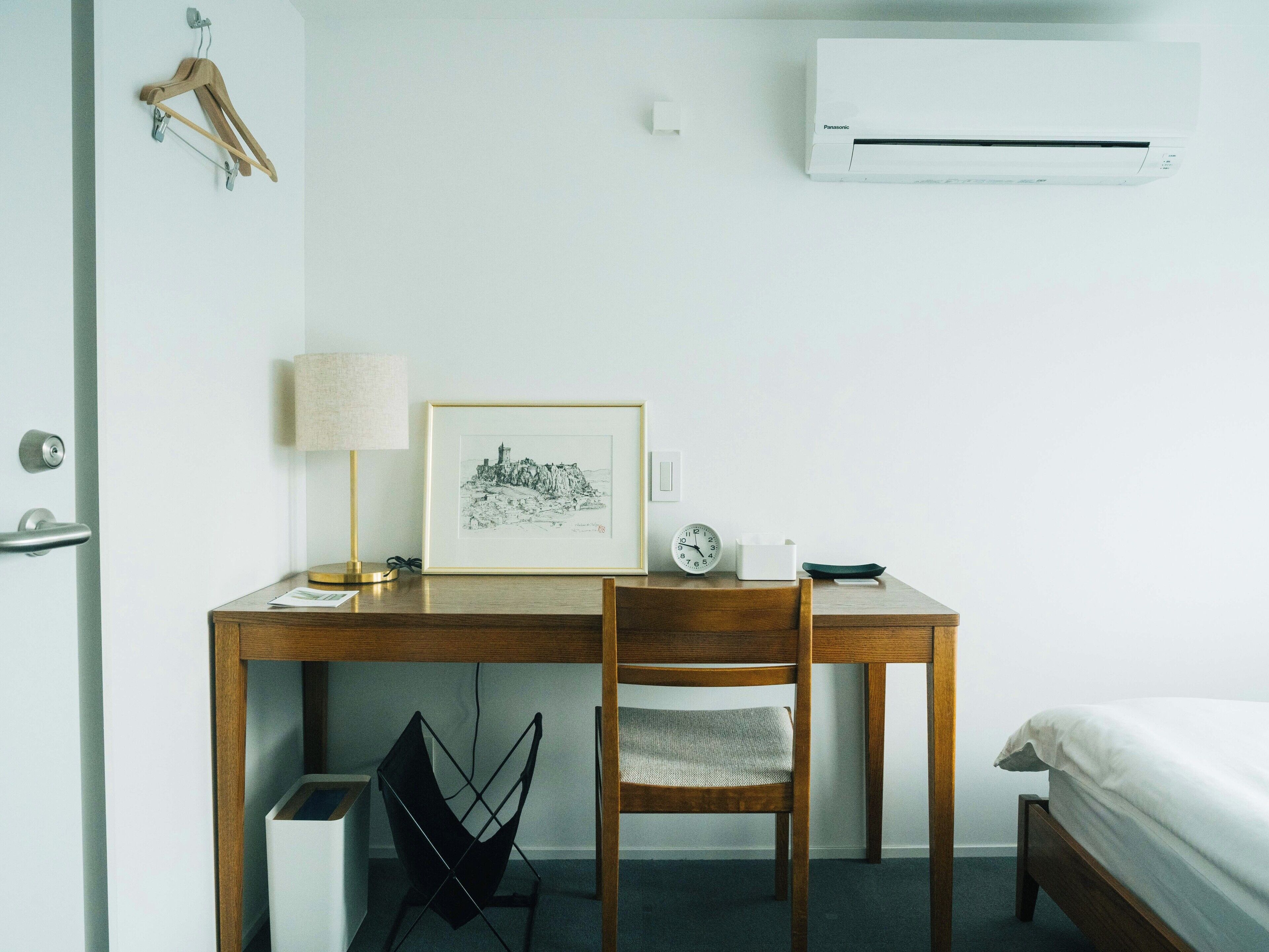 Ein schlicht eingerichtetes Hotelzimmer mit Holzschreibtisch, Stuhl, Bett, Wandklimaanlage, gerahmtem Bild, Nachttisch mit Lampe, einer Uhr und einem Papierkorb.