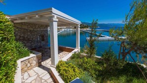 Zona junto a la piscina con pared de piedra, pérgola y vistas al mar y a las montañas.