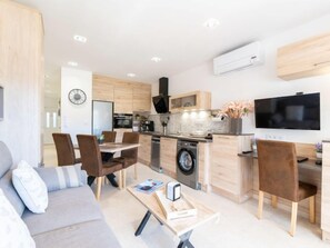 Un salon moderne avec un canapé, une table basse et un coin repas. La cuisine est équipée de meubles en bois, d’un lave-linge et d’un téléviseur.