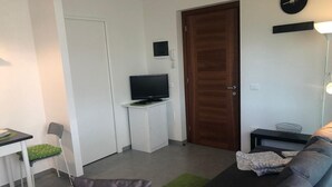 Una sala de estar con un sofá, un televisor sobre un mueble bajo, una puerta, una mesita con una silla y una lámpara.