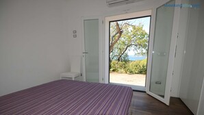 A bedroom with a bed, a chair, and a sliding glass door leading to an outdoor area with a view of trees and a body of water.