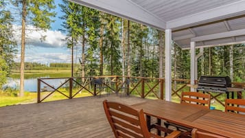Een overdekt terras buiten met een tafel en stoelen, met uitzicht op een meer en bos.