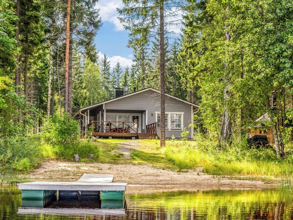 A cabin with a deck, surrounded by forest and a body of water.