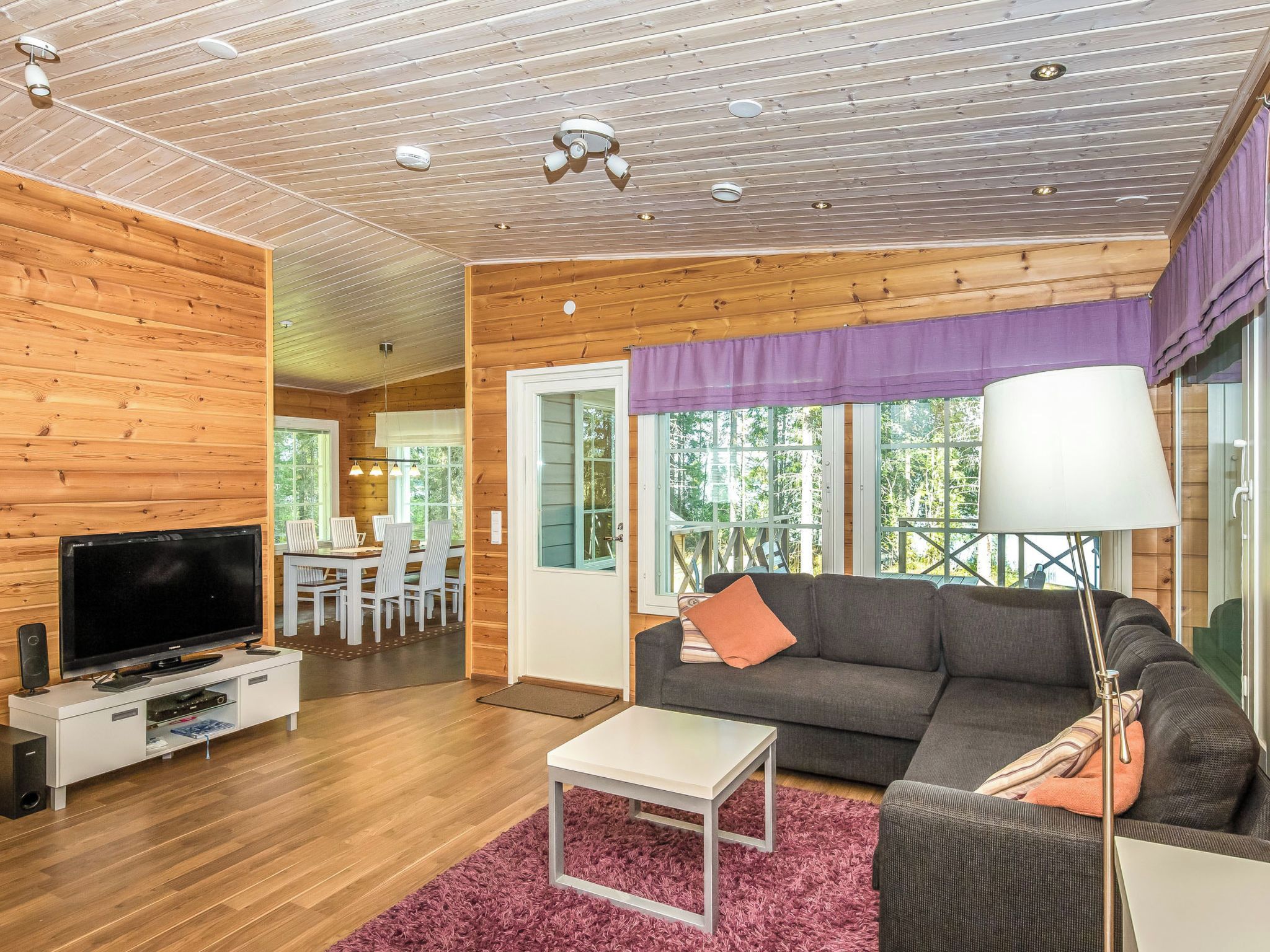 A modern living room with a flat-screen TV, a gray sofa, a white coffee table, and a purple window valance.