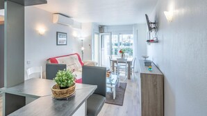 A modern living room with a dining area, a sofa, a table with a plant, and a kitchen area with a counter and a hanging light fixture. - Port Salins by Interhome (Empuriabrava)
