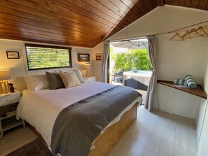 A bedroom with a wooden ceiling, a bed with a grey blanket, a nightstand with a lamp, a window with blinds, and a view of greenery outside.