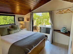 A bedroom with a bed, a nightstand, a lamp, a hot tub, and a view of greenery outside.