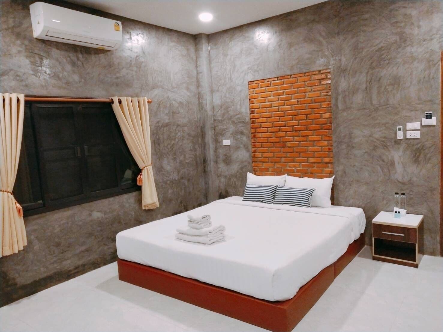 Un dormitorio con una cama grande, una pared de ladrillo decorativa, un equipo de aire acondicionado, una ventana con cortinas y una mesita de noche con una botella.