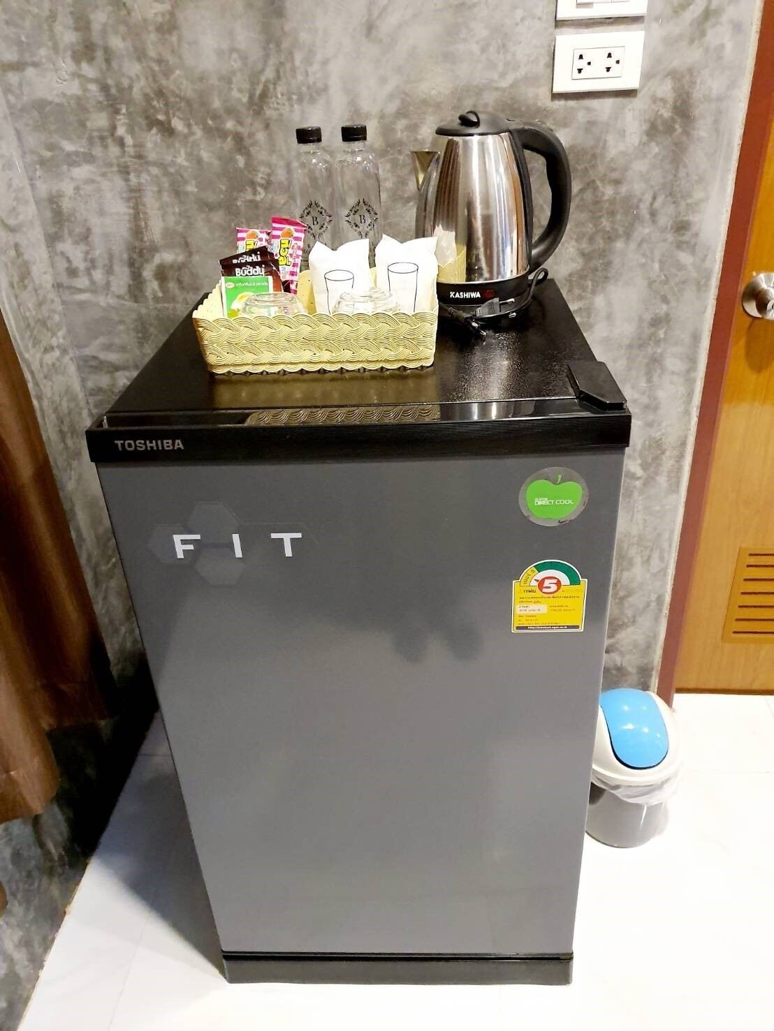 A compact refrigerator with a Toshiba logo, a direct cool sticker, and a few items on top including a bottle, a box, and a kettle.