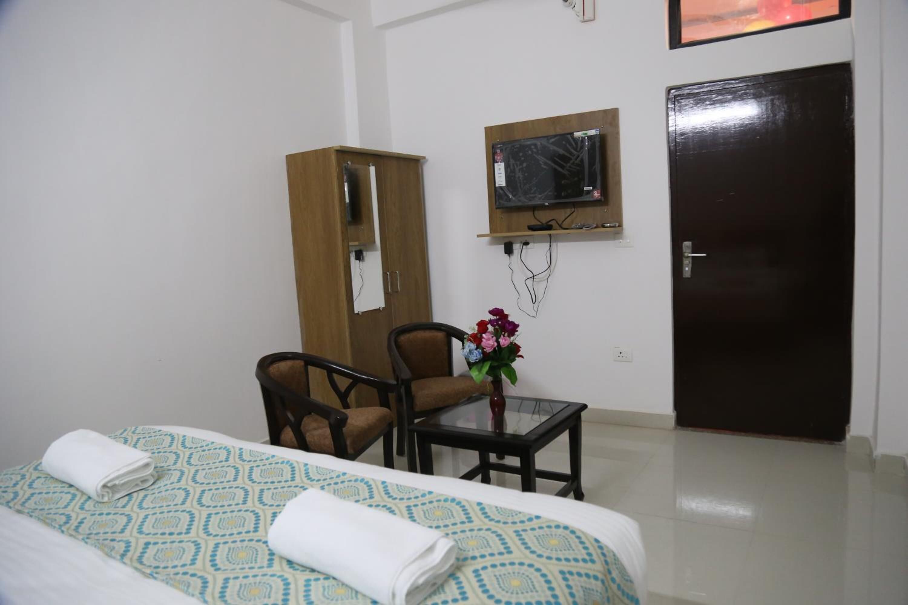 A hotel room with two beds, a wooden wardrobe, a television mounted on the wall, a small table with a vase of flowers, and a door.
