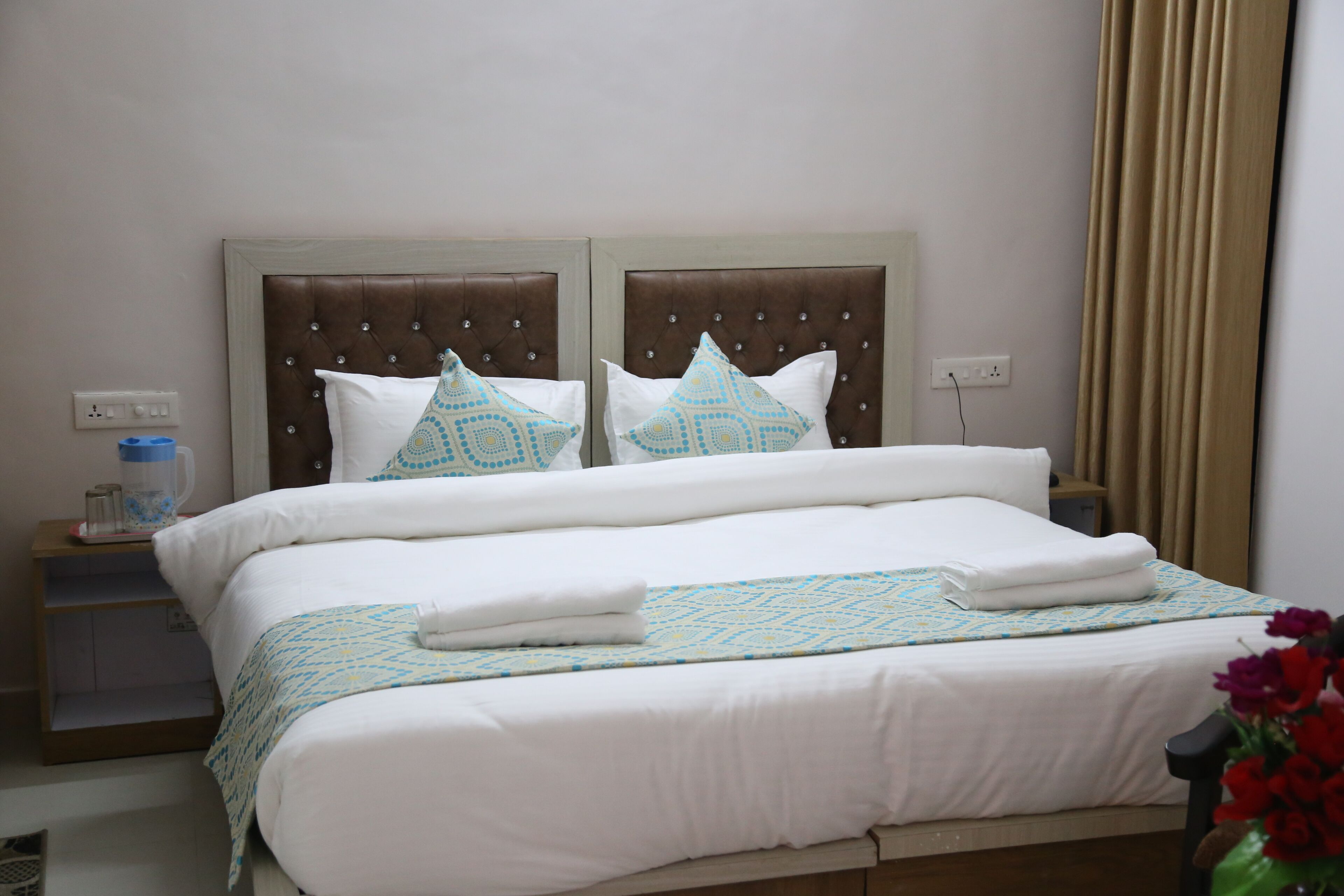 Una cama bien hecha con una colcha estampada, dos almohadas con diseños geométricos y un jarrón con flores rojas a un lado.