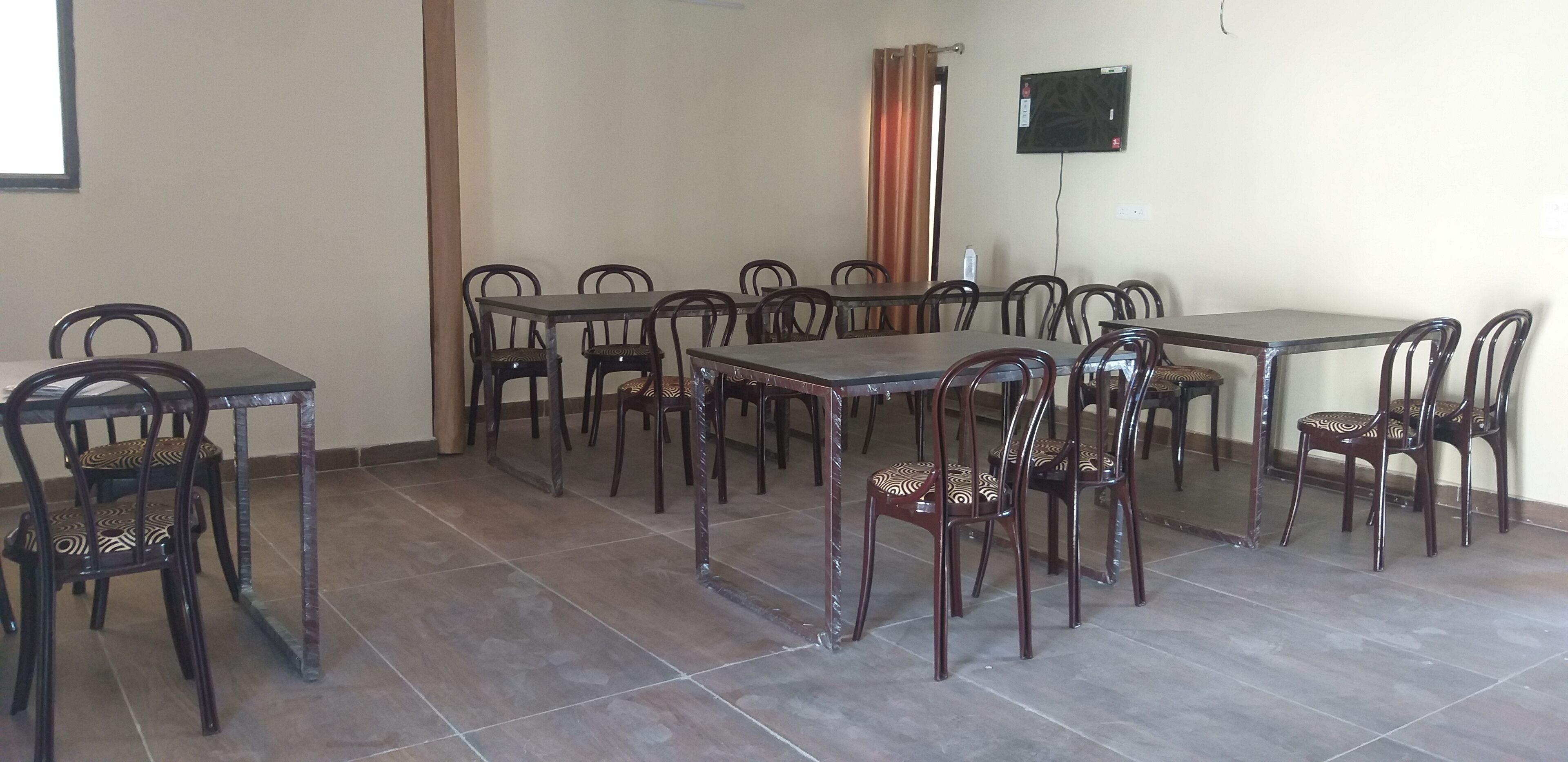 A dining area with multiple tables and chairs, a television mounted on the wall, and a curtain partially drawn.