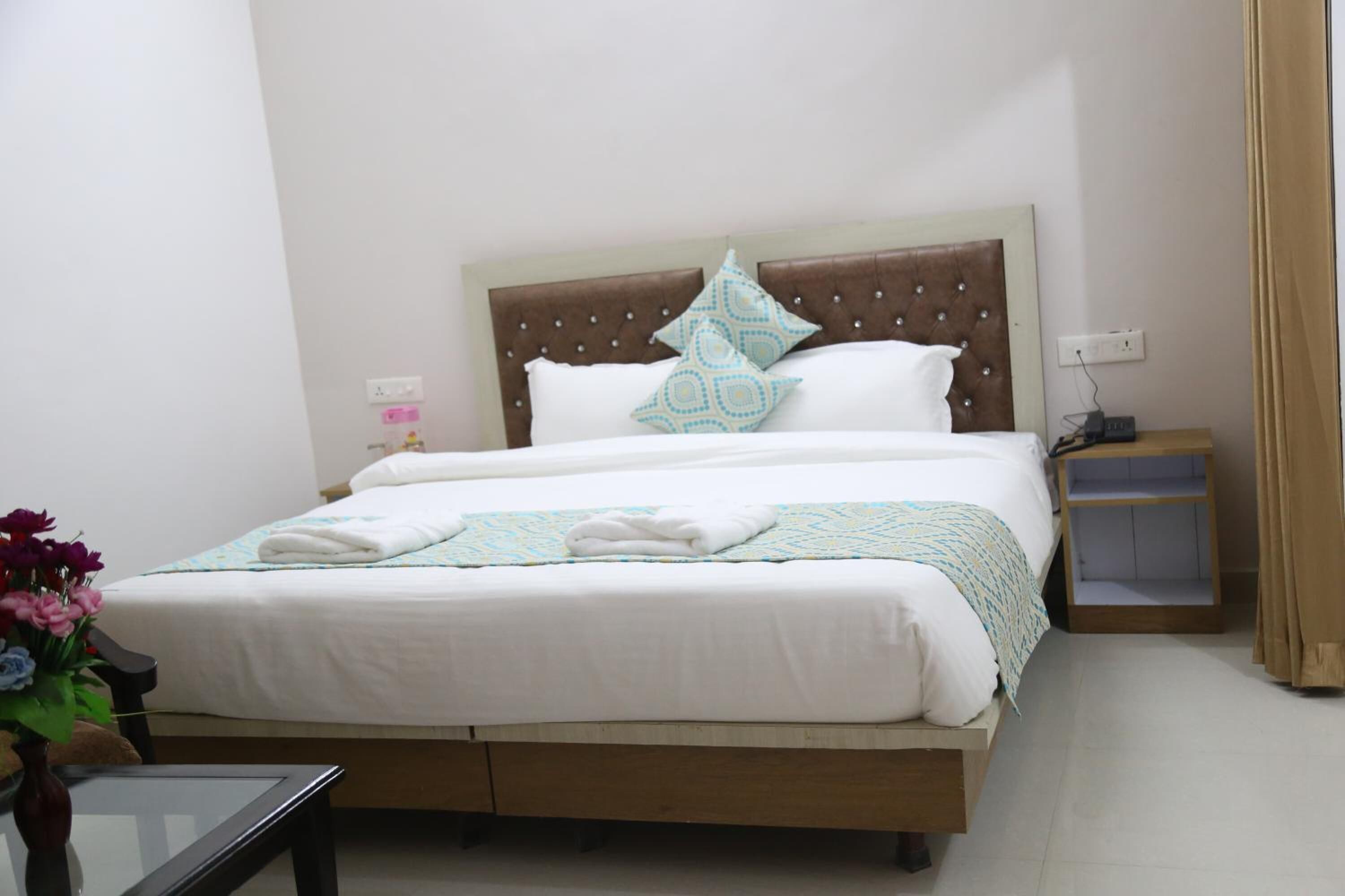 A neatly made bed with white linens and a patterned pillow, a bedside table with a lamp, and a small wooden nightstand with a phone charger.