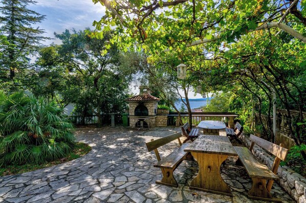 Un patio empedrado con bancos de madera y una chimenea de piedra, rodeado de abundante vegetación y con vistas a un cuerpo de agua.