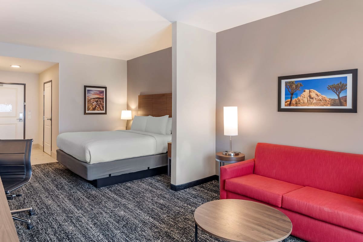 A hotel room with a bed, a red sofa, a round coffee table, a desk with a chair, and two framed pictures on the walls.