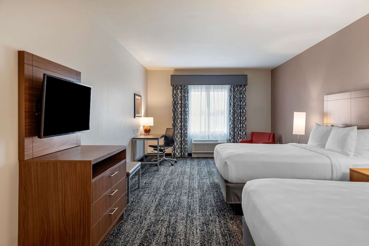 A hotel room with two beds, a desk with a chair, a TV mounted on a wooden cabinet, and a window with curtains.